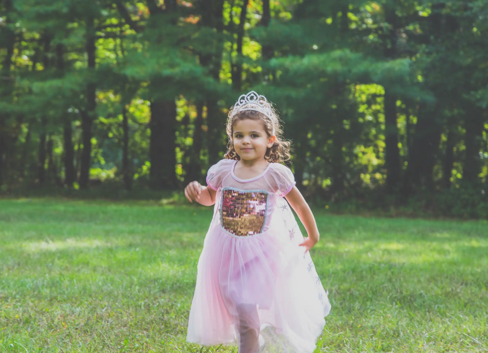 Little girl in a pink princess dress and tiara running across a sunlit lawn