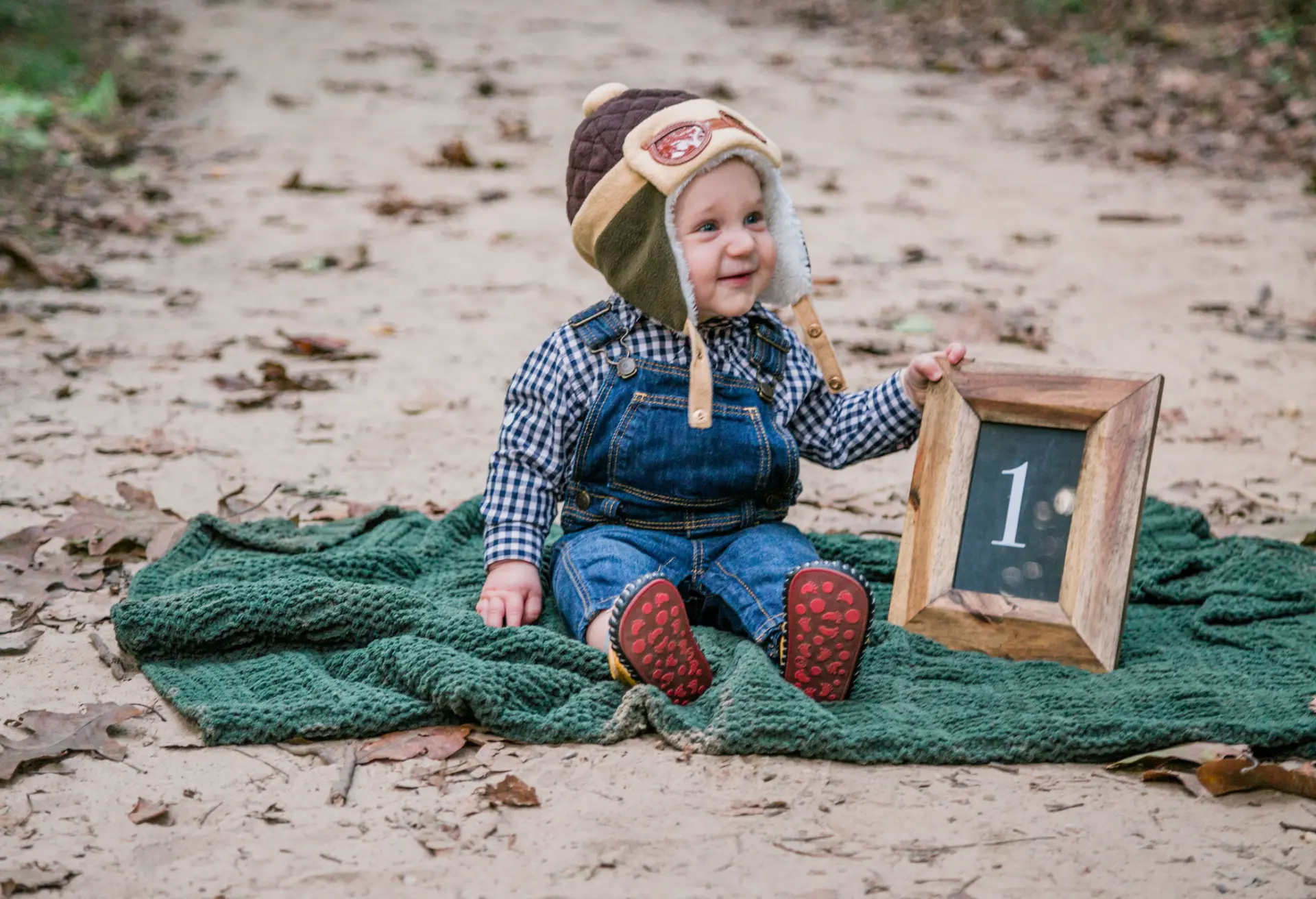 One-year-old in overalls and an aviator hat with a chalkboard number one
