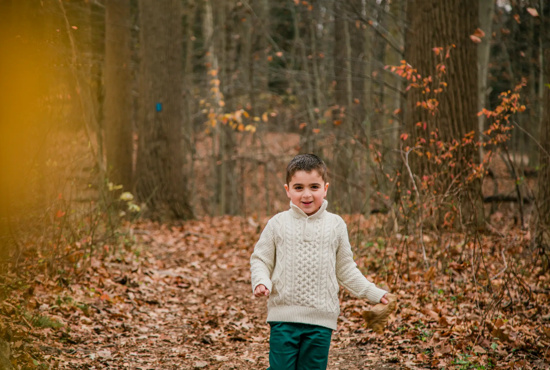 Boy in a cream cable-knit sweater on a leaf-covered woodland trail