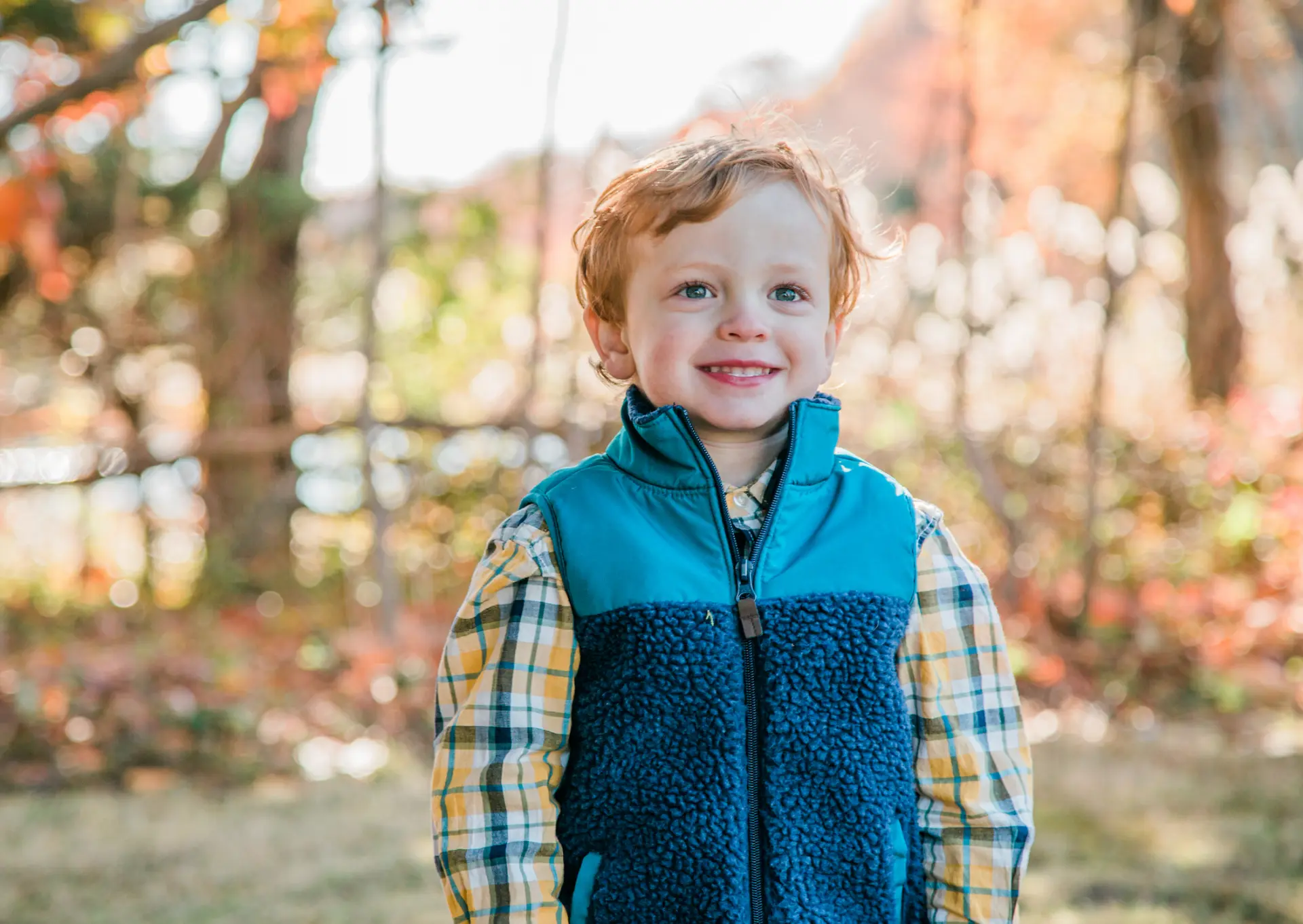 Red-haired toddler boy in a teal fleece vest in autumn light