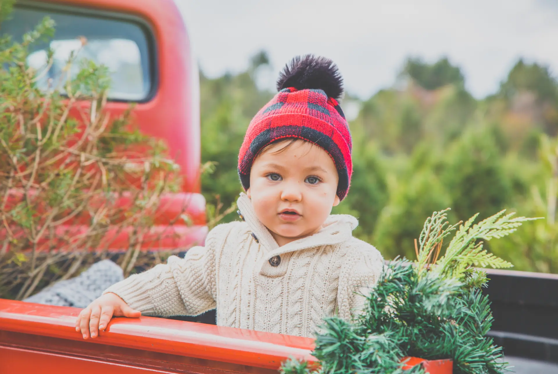 Baby in a plaid pom hat in a red vintage truck at a tree farm