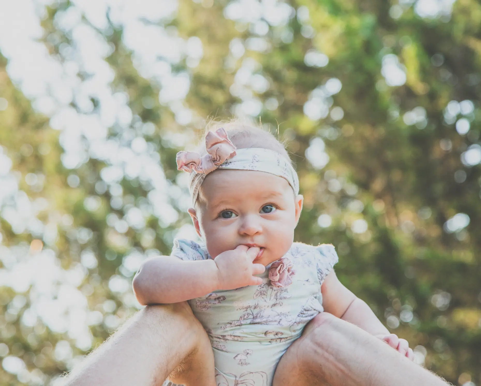 Baby girl in a floral romper and bow headband held up toward the sunlit trees