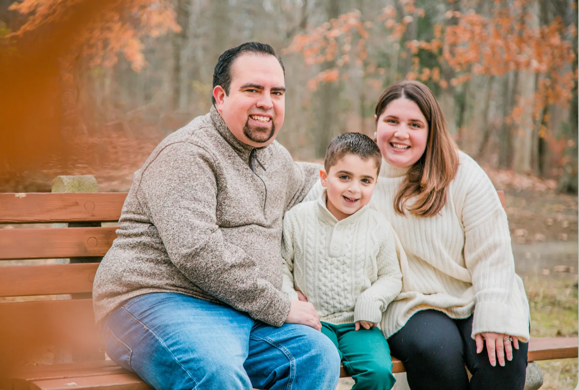 Family of three smiling on a park bench framed by orange autumn leaves